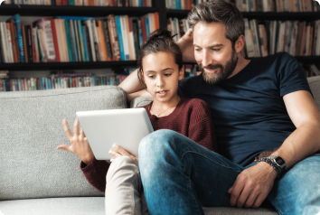 An adoptive dad sits on a couch with his daughter as they look at a tablet and consider searching for her birth family.