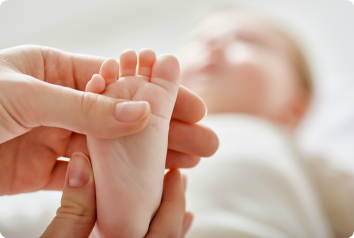 A close up shot of the foot of an infant who was adopted as she is at the doctors office. The rest of her body is visible but blurred in the background.