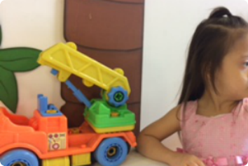 A photo of Y sitting at a table. There is a toy crane made of primary colors on the table and she is looking to the right. The photo only shows the side of her face to protect her privacy. She is waiting in South America for an adoptive family.