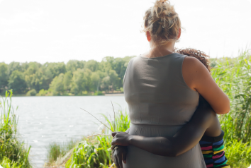 A photo of the backs of a mother and her preteen daughter, who was adopted from foster care, as they hug and look over the lake.