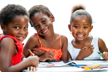 Three elementary-age black girls smile at the camera as they sit around a table drawing.