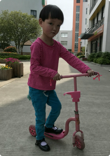 A photo of YY playing on a scooter in a paved area. She is looking shyly at the camera.
