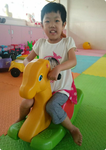 XW is pictured riding a yellow rocking horse within the orphanage. She has a sweet smile and a pixie hair cut.