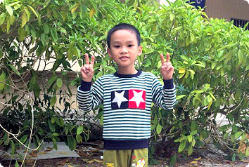 Nine year old boy waiting for adoption in Asia shown smiling and holding up peace signs in front of a bush