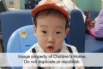 A young Asian boy waiting for adoption looks at the camera, wearing an orange hat.