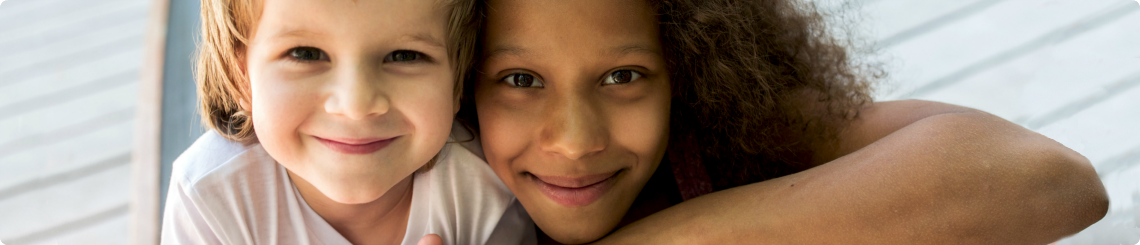 A sister and brother smile at the camera. The sister is a preteen African American girl, the brother is a toddler white boy.