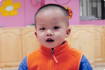 A little boy in an orange vest looks at he camera. He waits for an adoptive family.