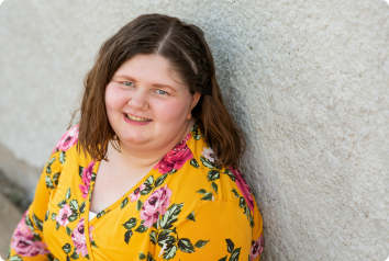 Sherakey, a teen waiting in foster care for adoption, leans against a white wall and smiles in a bright yellow dress.