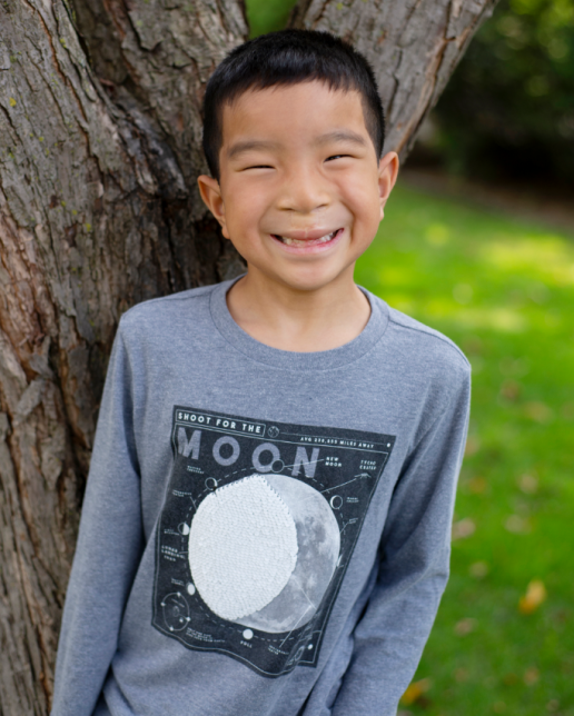 School-age Asian boy smiles wearing a grey long sleeve shirt in front of a tree.