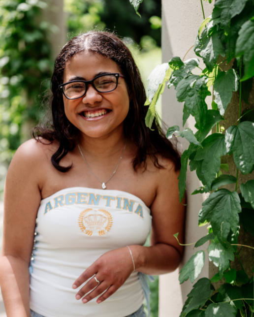 Trinity wearing a white top reading "Argentina" and smiling by an ivy-covered wall on a sunny day