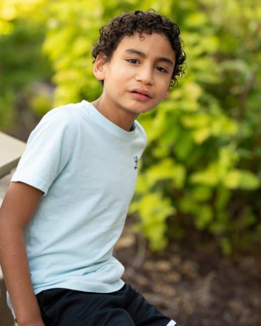 Portrait of JJ wearing a light blue tshirt in front of green leaves on a sunny summer day