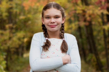 Saphira wearing a white shirt with braided pigtails standing in front of fall colors with her arms crossed.
