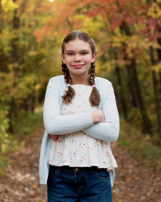 Saphira wearing a white shirt with braided pigtails standing in front of fall colors with her arms crossed.